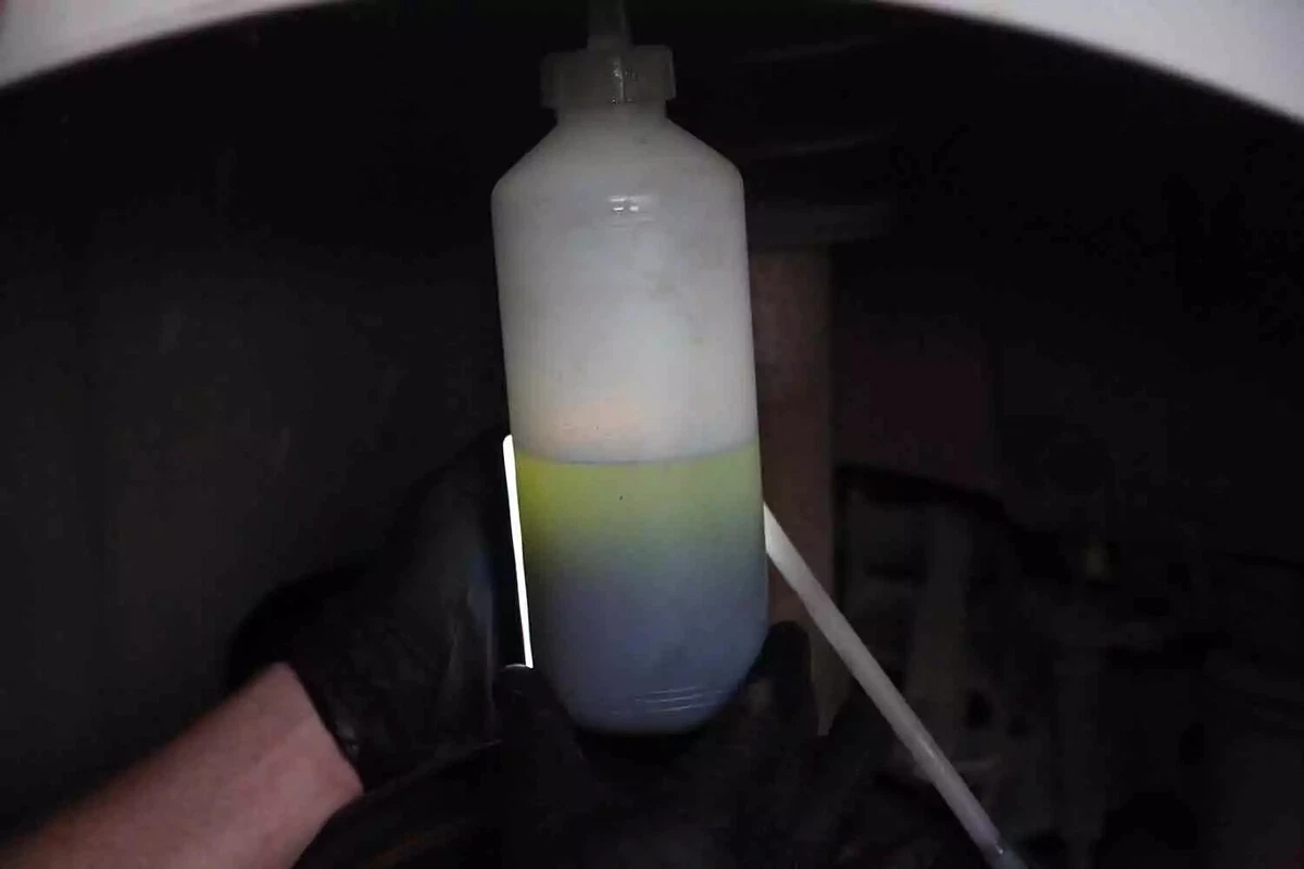 A mechanic wearing a black glove holds the catch bottle of a brake bleed kit as it fills with dirty brake fluid.