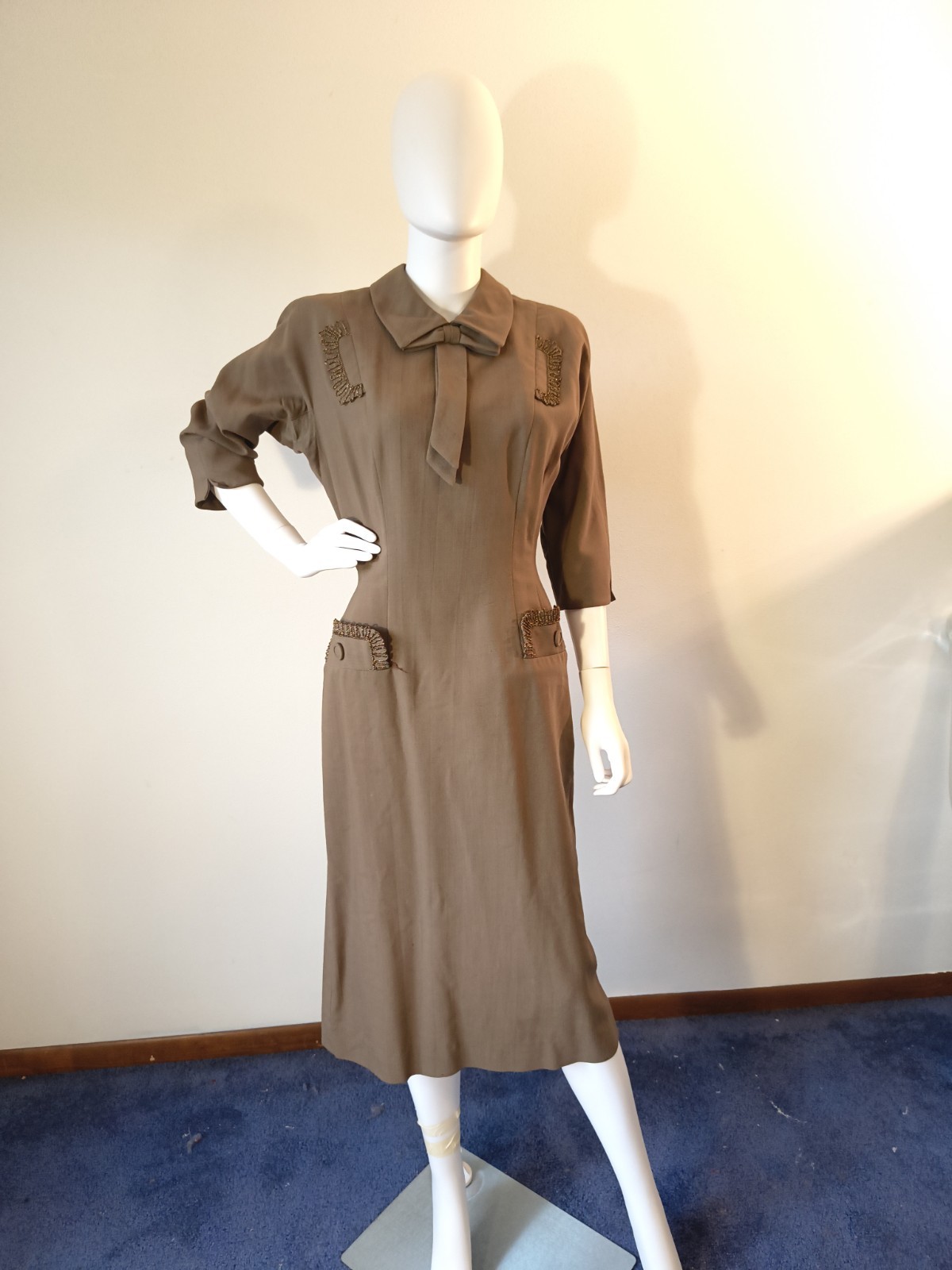 Olive-brown 1950s wool dress with beaded trim on the collar and pockets, on a dress form.
