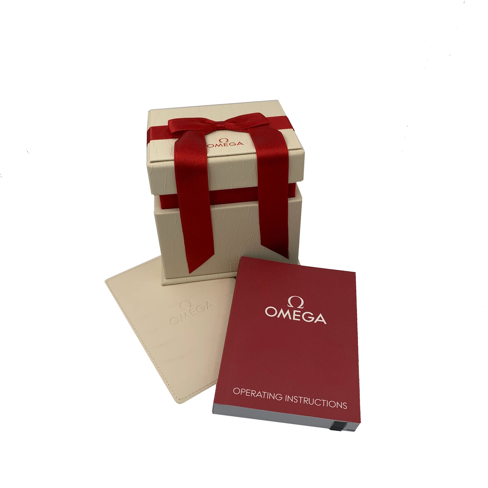 An image of an OMEGA wristwatch display model for women, shown in its packaging. The watch is not visible, but the beige box with a red ribbon and OMEGA logo is centered in the frame. In front of the box are an OMEGA operating instructions booklet and a beige document holder, positioned slightly to the right. The image is taken from a slightly elevated angle at a close distance.