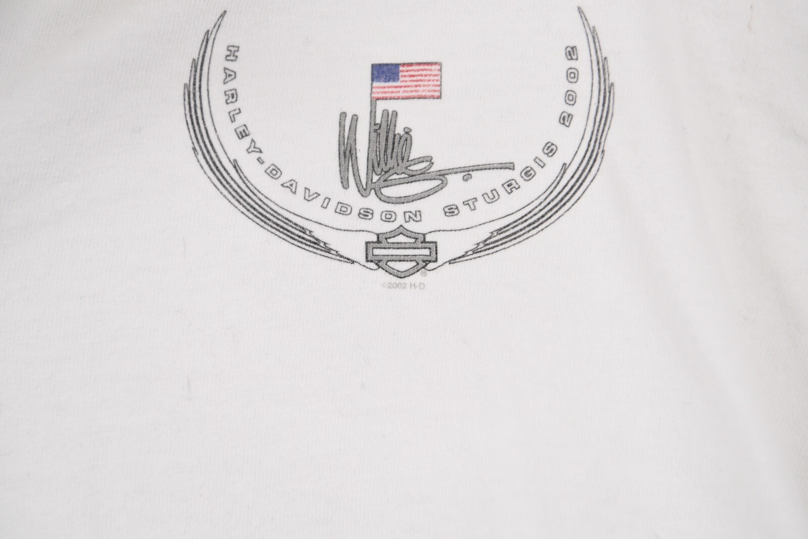 White vintage Harley-Davidson Sturgis 2002 polo shirt with black embroidered winged logo and red, white, and blue USA flag.