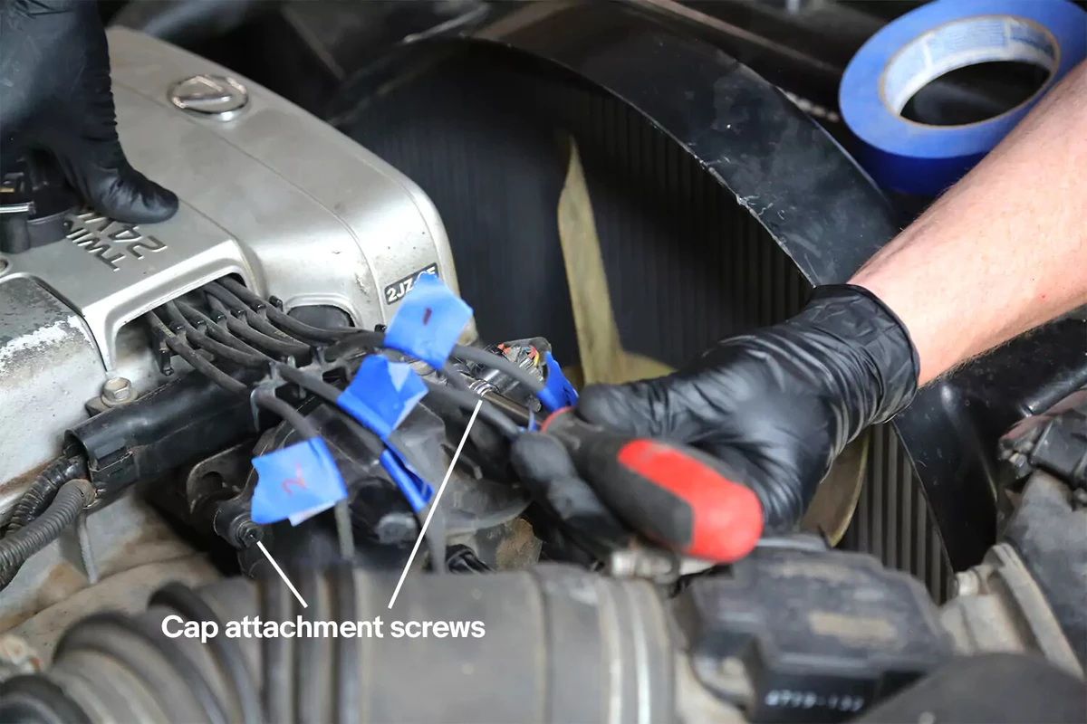 A mechanic wearing black gloves uses a screwdriver to remove cap fasteners.