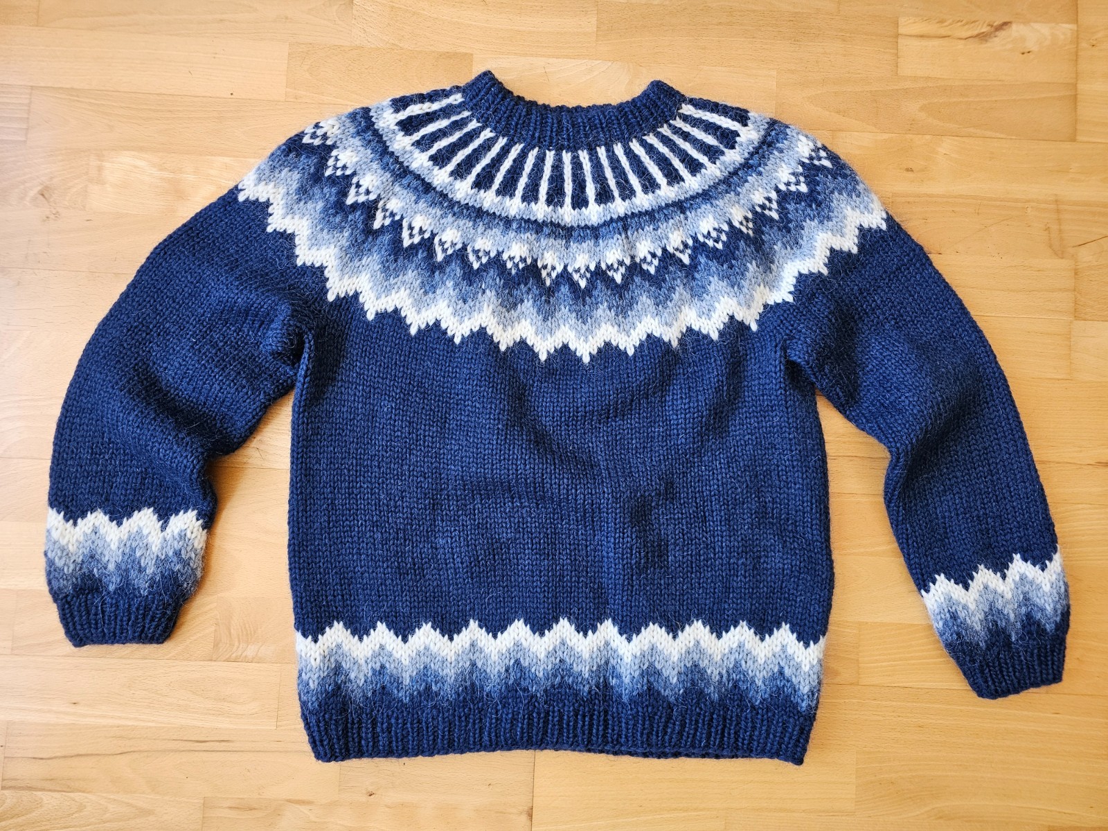 Navy and white hand-knit Icelandic lopapeysa wool sweater with yoke pattern, laid flat on a wood floor.