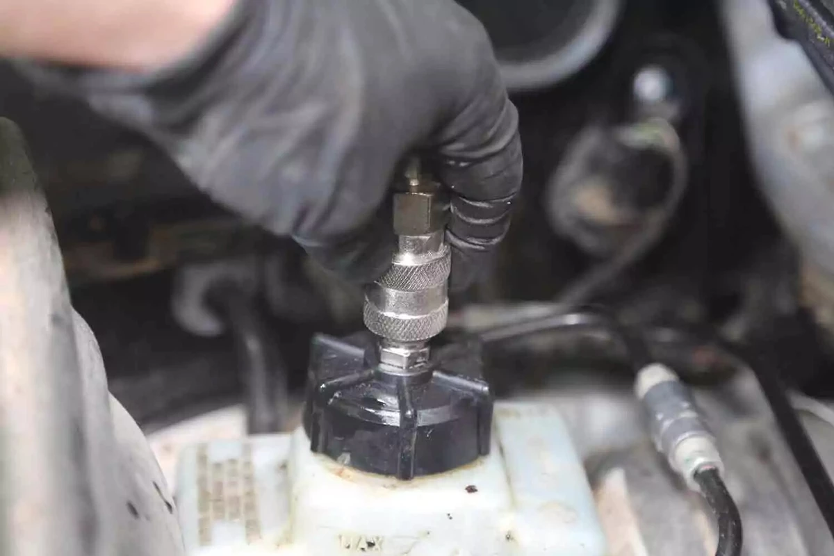 A mechanic wearing a black glove disconnects the hose from the cap of a vehicle's brake fluid reservoir.