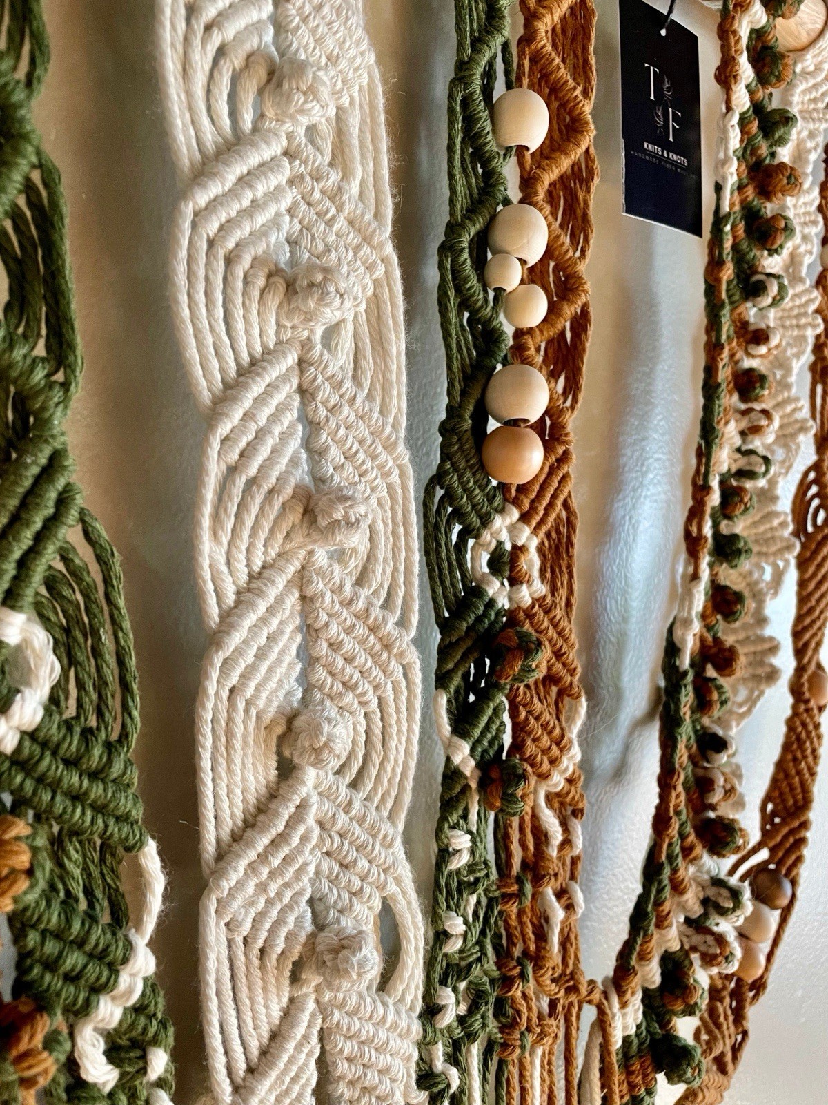 Large white macrame wall hanging with intricate braided patterns and wooden beads, displayed among similar pieces.