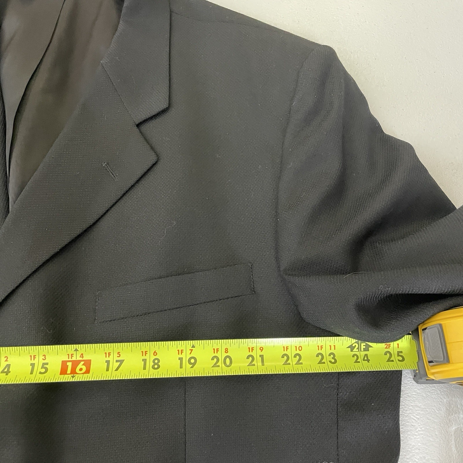 Black solid three-button blazer jacket with notch lapels, front flap pockets, and yellow measuring tape across the chest.