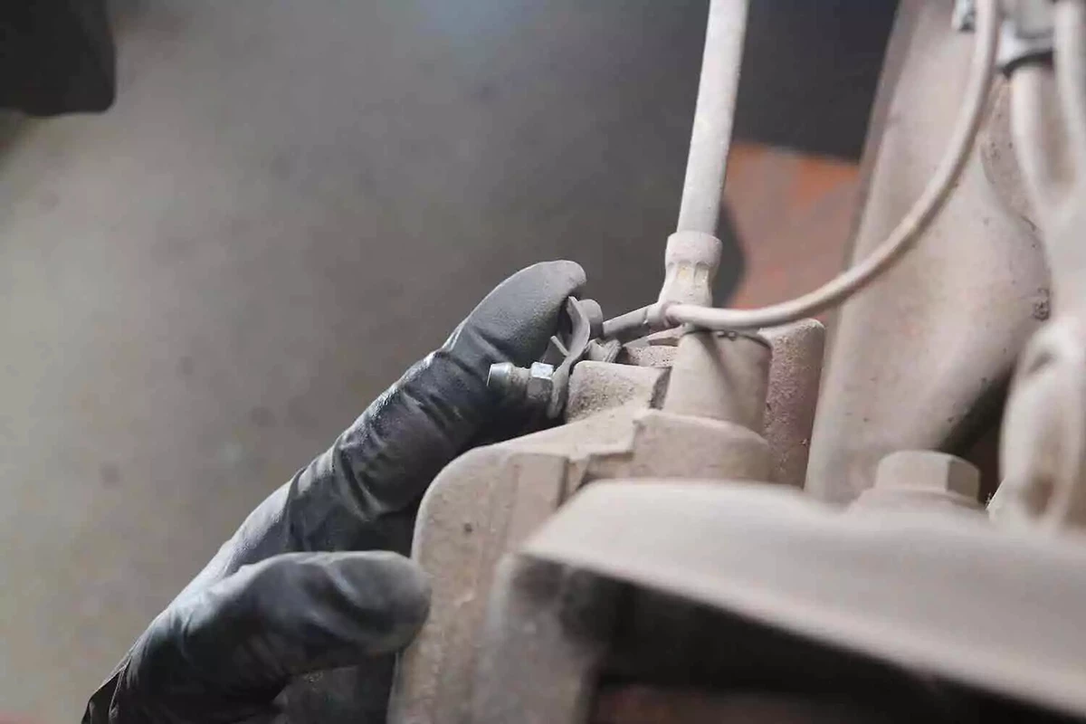 A mechanic wearing a black glove points to the vehicle's brake bleeder.