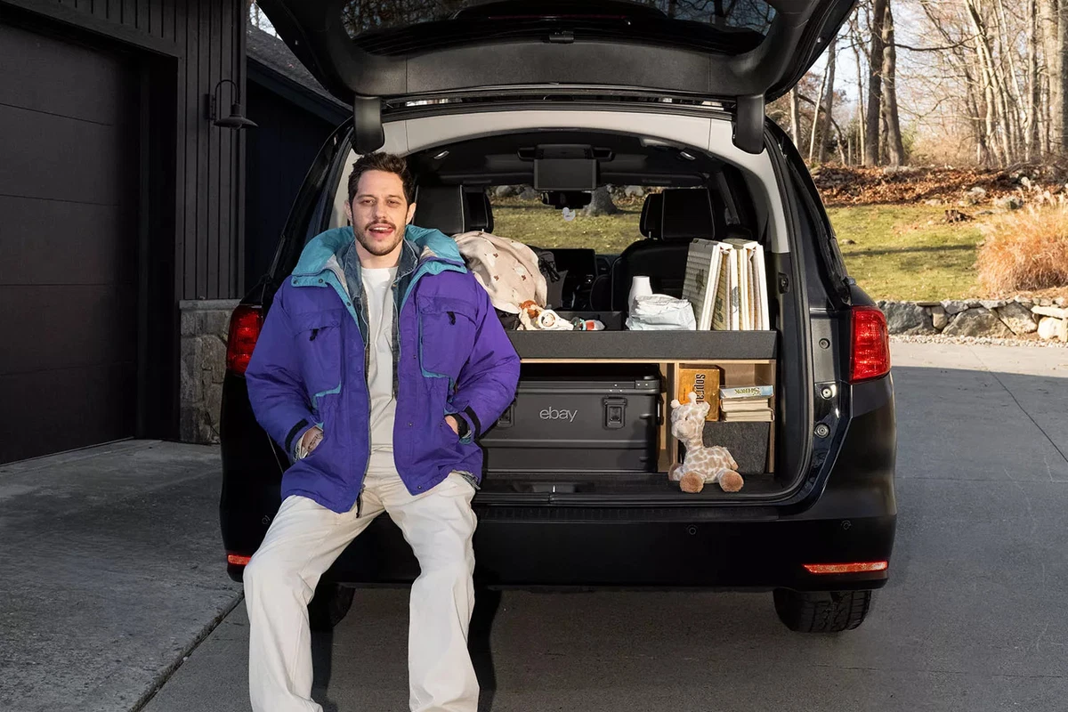 Actor and comedian Pete Davidson sits on the bumper of a modified minivan with its rear gate open.