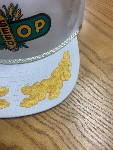 White vintage trucker hat with green and yellow Coop Seed logo, featuring embroidered cornfields and trees on the front panel.