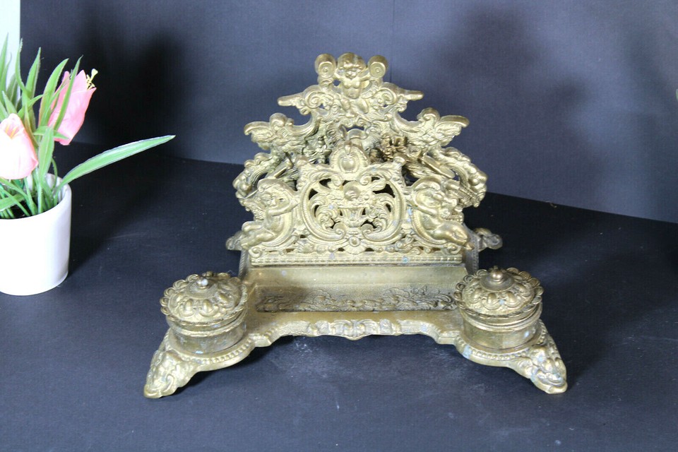 An antique brass inkwell held aloft by putti cherubs — a genuinely museum-worthy sculptural desk piece dripping with old-world drama.