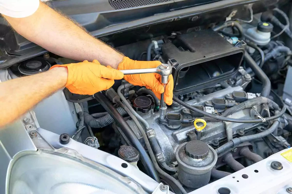 A mechanic wearing orange gloves uses a socket wrench to remove an ignition coil in order to replace a spark plug.