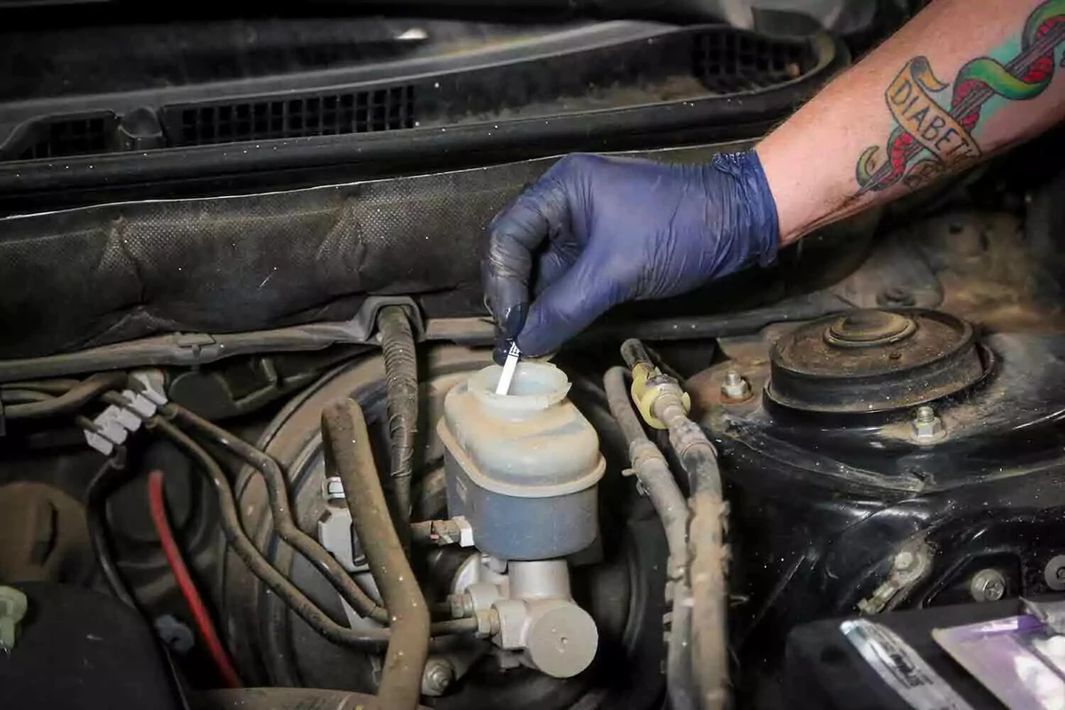 A mechanic wearing a blue nitrile glove inserts a brake test strip into the master cylinder’s fluid reservoir.