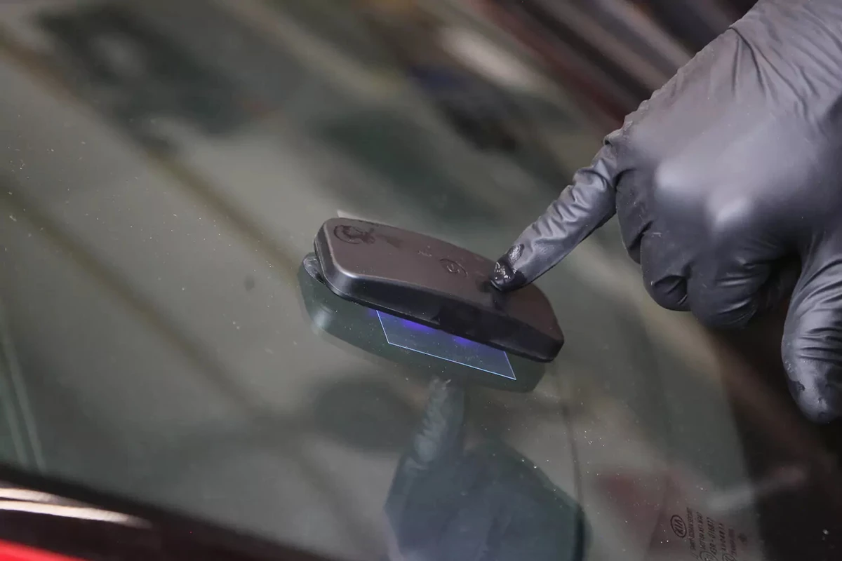 A mechanic wearing a black glove holds a UV light over the repaired part of a windshield.