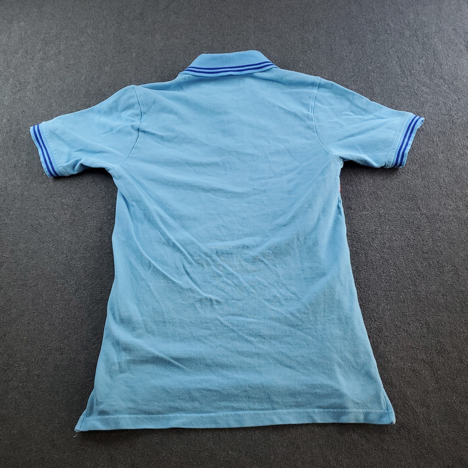 Light blue boys’ polo shirt with white sleeve trim, blue shirt buttons, and “Aston Martin Racing Team” logo on back.