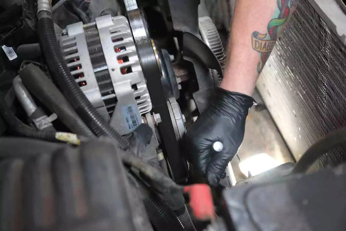 A mechanic wearing a black glove uses a ratchet to release the serpentine belt tension.