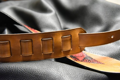 Brown leather guitar strap with dual loops and inlaid tuscon shell design, displayed on black fabric background.
