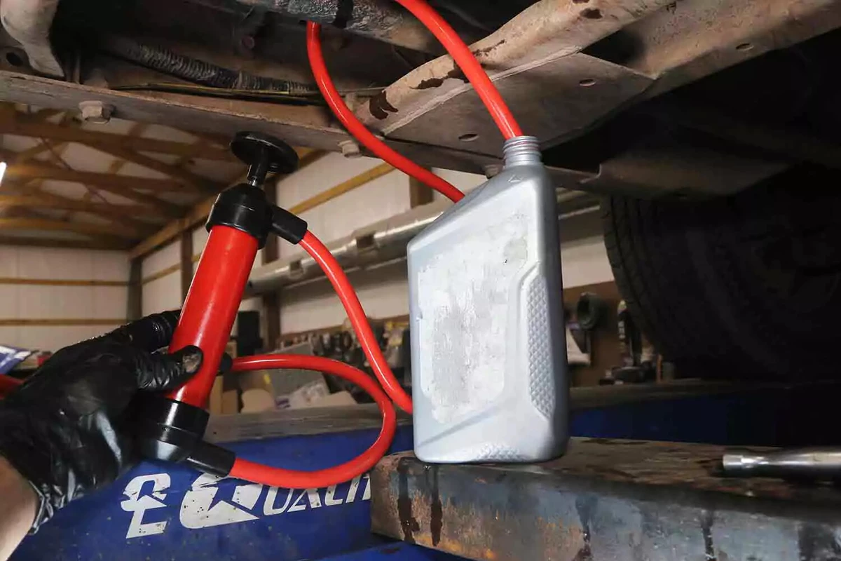 A mechanic wearing a black glove uses a hand-operated pump to add transfer case fluid to a vehicle.