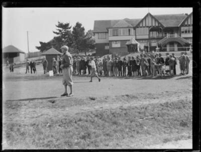 Golfer T. Howard hitting a ball in front of crowd by a club house,- Old Photo