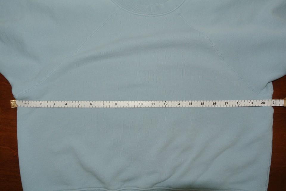 Light blue French-terry sweatshirt laid flat with a physical measuring tape across its chest