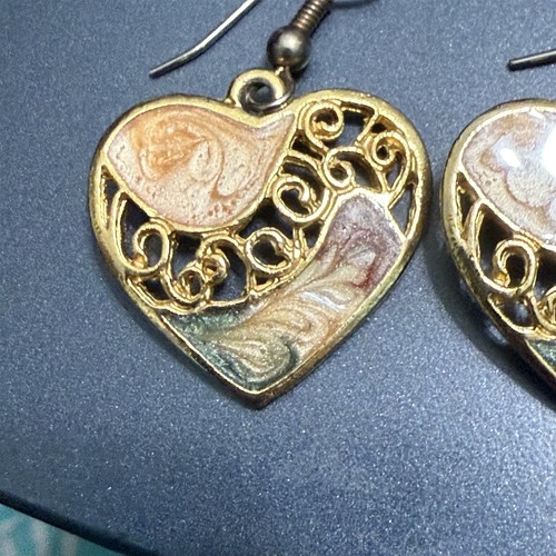 Gold-tone dangle drop earrings with filigree swirl design and round enamel heart pendants on a dark blue background.
