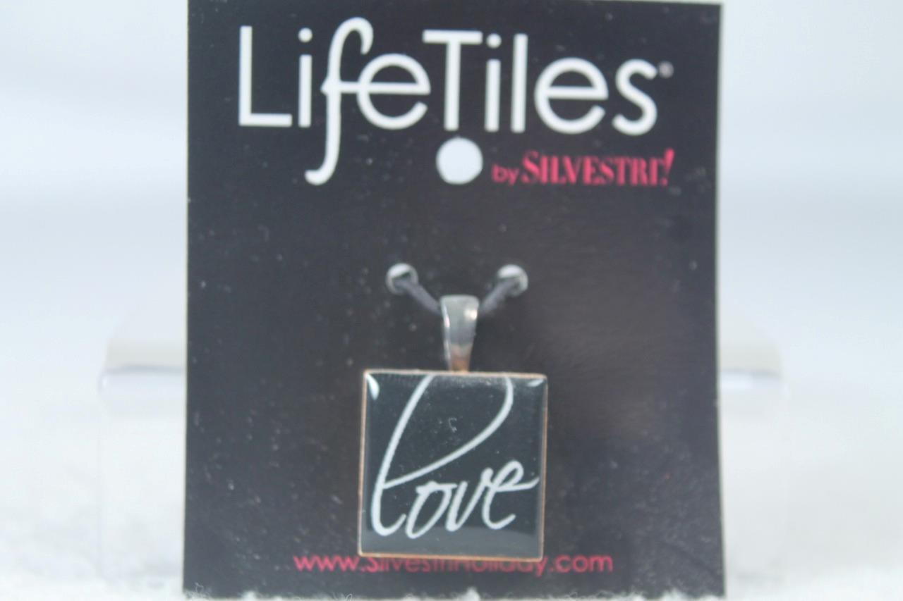 Silver metal pendant with square glass tile displaying white “LOVE” text, hanging from black card labeled “Life Tiles.”