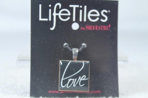 Silver metal pendant with square glass tile displaying white “LOVE” text, hanging from black card labeled “Life Tiles.”