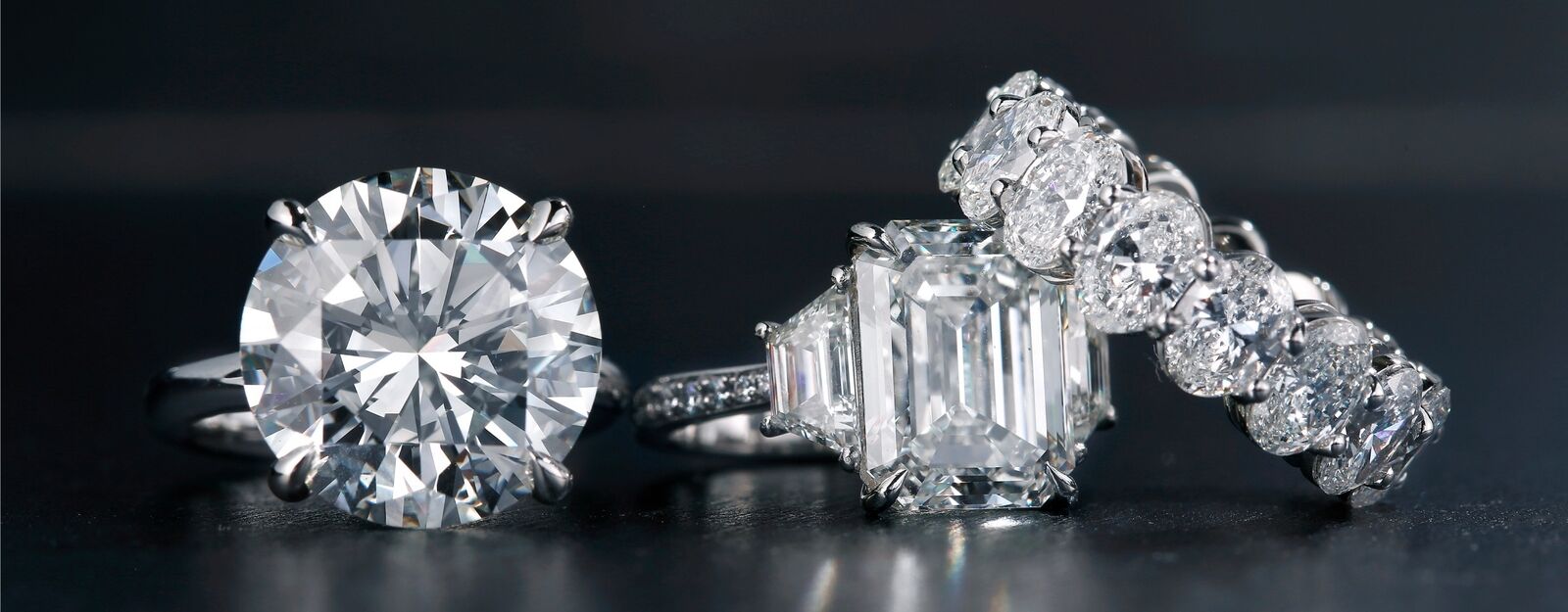 Nahaufnahme von drei funkelnden Diamantringen mit unterschiedlichen Schliffen auf dunklem Hintergrund.