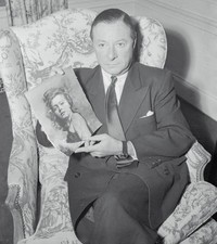 Comedian George Jessel looks serious as sits holding a photo of f- 1956 Photo