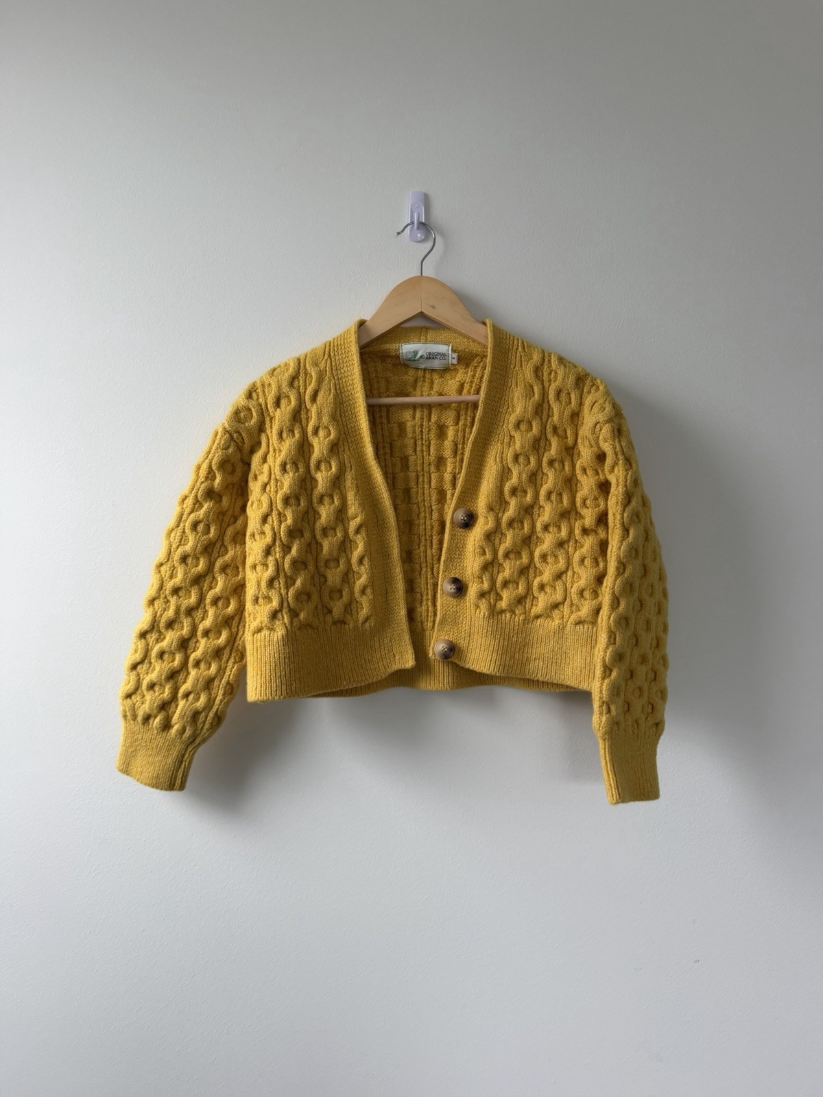 Mustard yellow chunky cable-knit cropped cardigan with wood buttons, shown on a hanger.