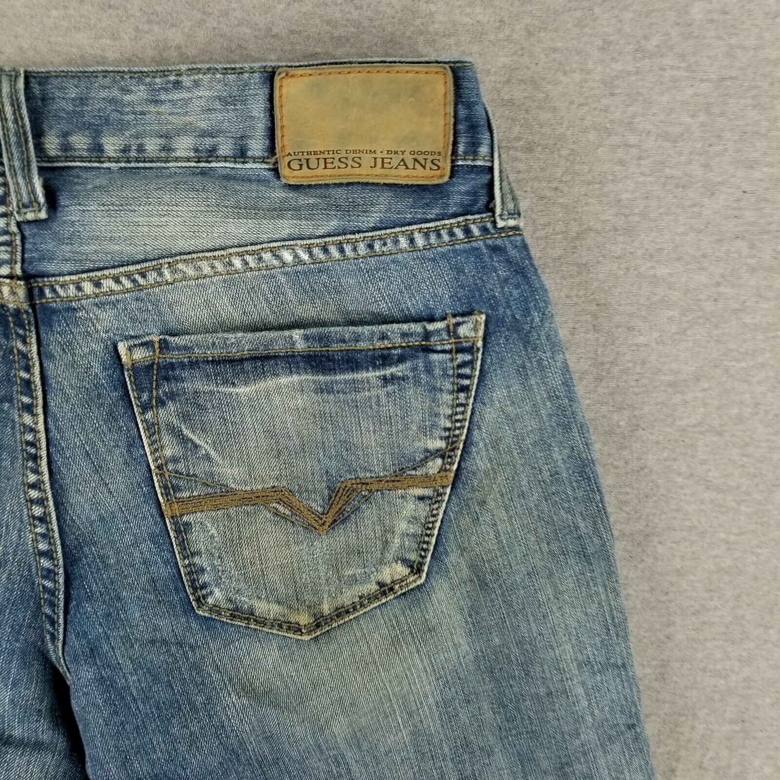 Back view of vintage blue GUESS jeans with asymmetrical button fly, tan leather patch, and visible wear marks on fabric.