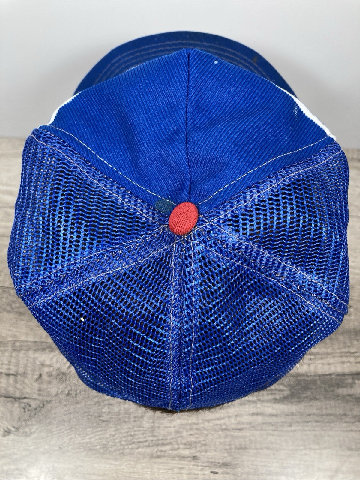 Blue and white snapback trucker hat with gold stitching and red button, shown from the back on a wooden surface.