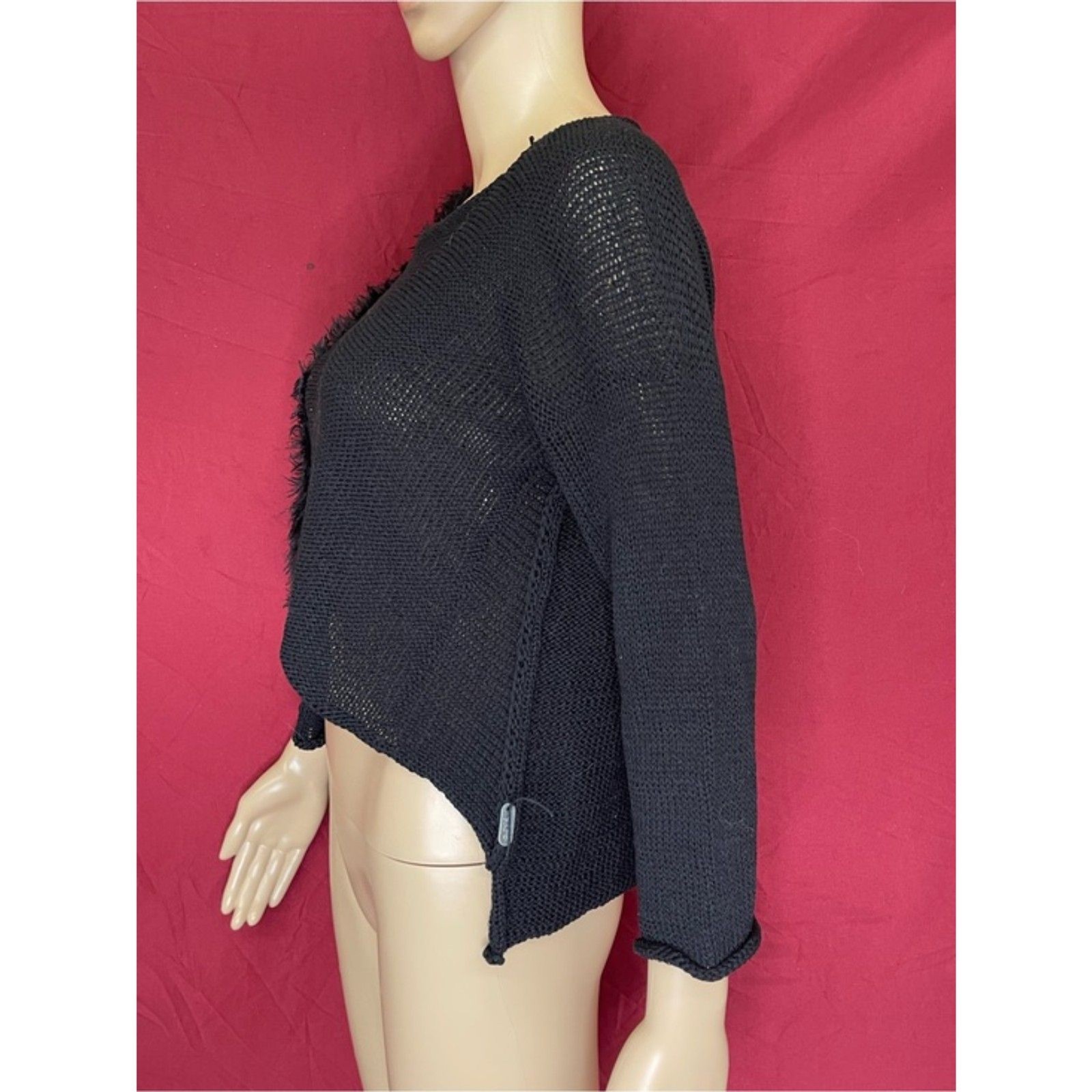 Black asymmetric crop sweater with long sleeves and loose weave, displayed on mannequin against red background.