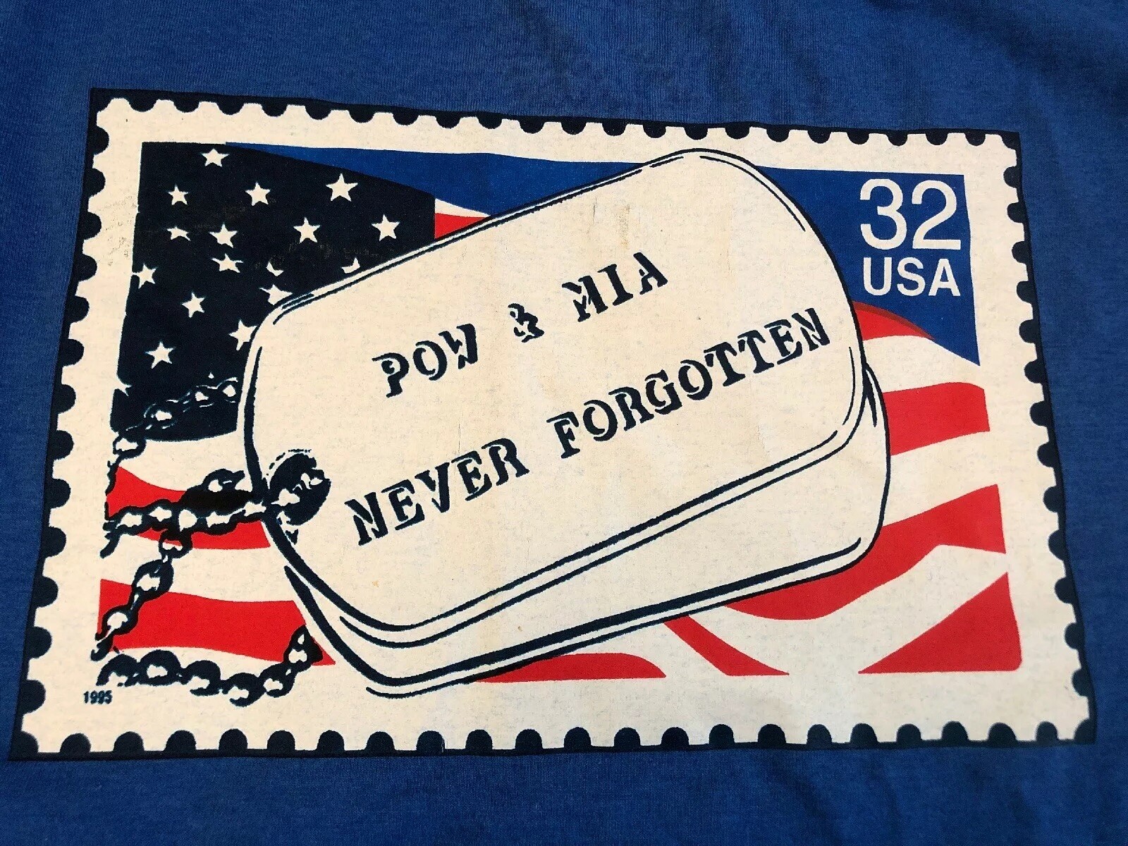 Blue EUC vintage 1995 T-shirt with U.S. flag design and text “POW & MIA Never Forgotten” on white dog tag.