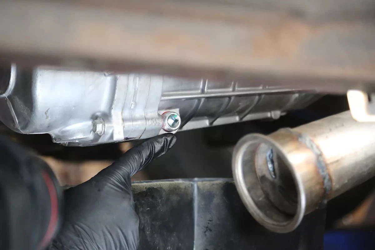 A mechanic wearing a black glove points to the drain plug of a manual transmission.
