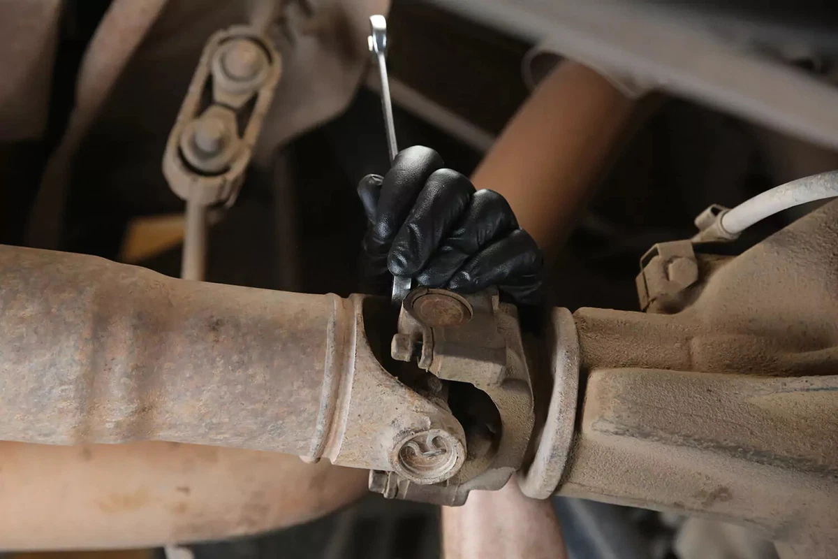 A mechanic wearing black gloves removes the bolts holding the caps of the U-joint in place.
