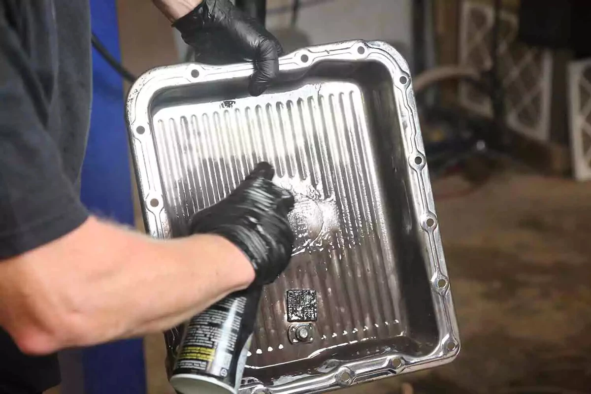 A mechanic wearing black gloves sprays the inside of a transmission pan with brake cleaner.