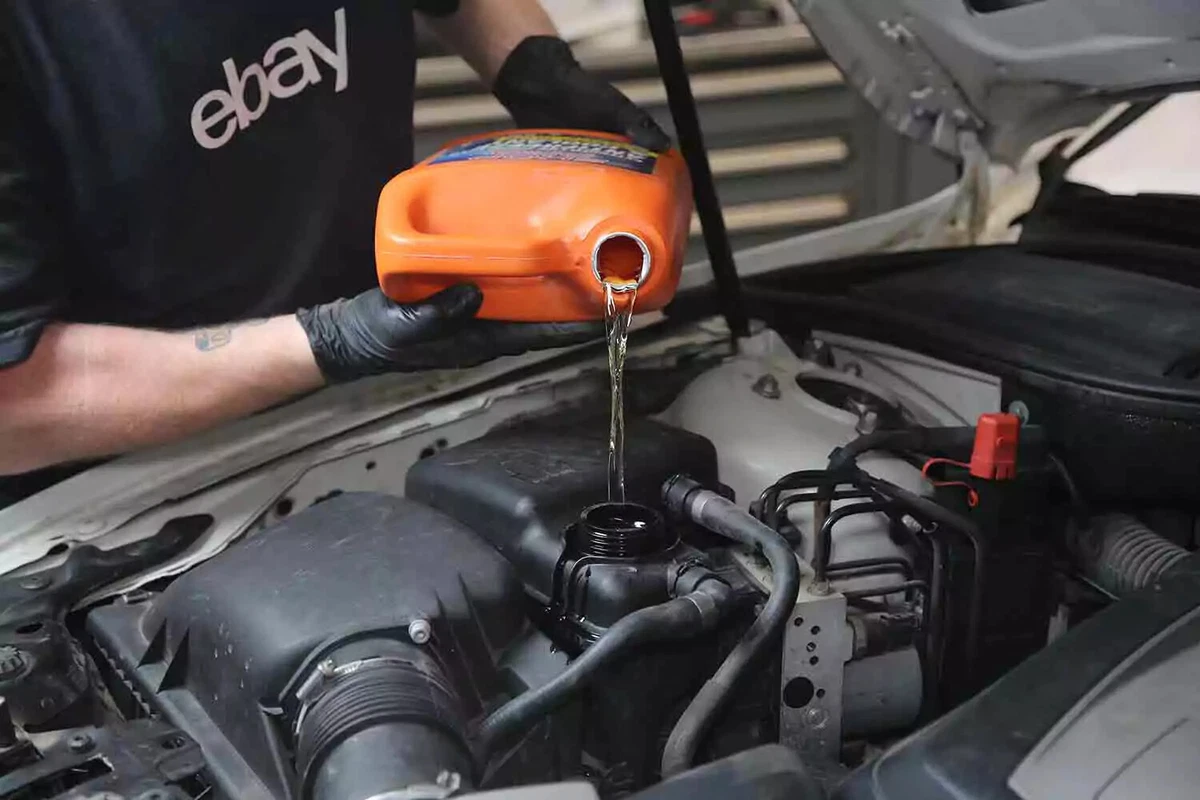 A mechanic pours new coolant from an orange container into a vehicle's coolant expansion tank.