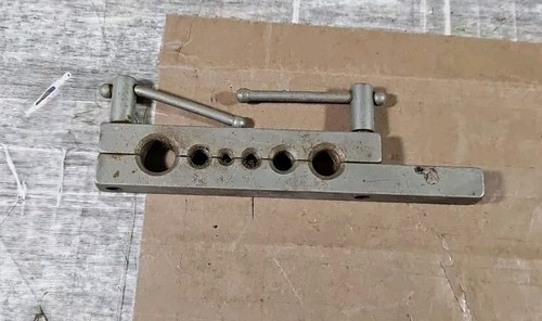 Vintage metal pipe and tube flaring tool with six circular holes and adjustable clamp, lying on a brown cardboard surface.