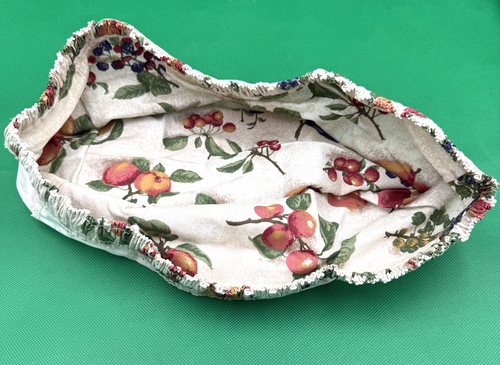 White fabric basket with red and green fruit print, blue edges, and visible stitching, resting on a green background.