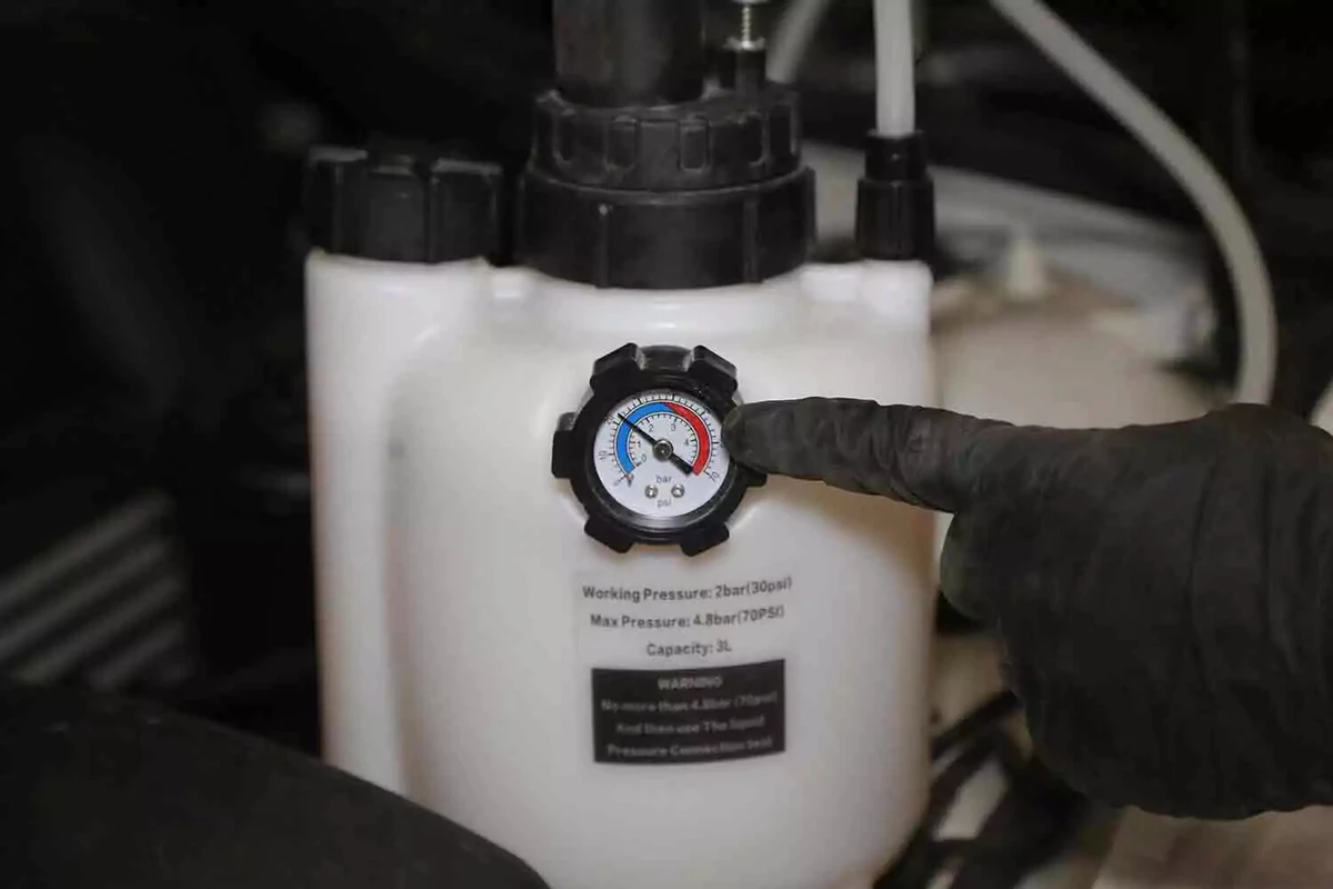 A mechanic wearing a black glove points to the PSI indicator at the front of a brake bleeder.