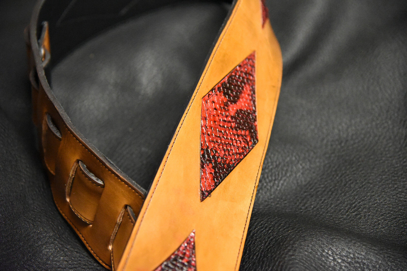 Brown leather guitar strap with red diamond inlay made of tan fabric, featuring two fastening holes and black strap.