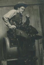 COWBOY IN HAT AND VEST WITH GUN IN HOLSTERS, POSED ON SADDLE ON OIL DRUM.