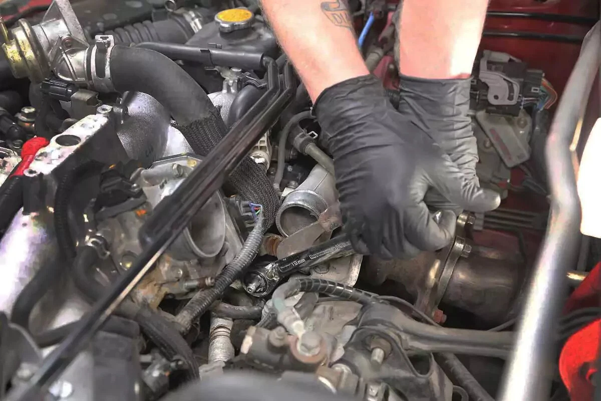 A mechanic wearing black gloves uses a socket wrench to loosen the PCV valve.