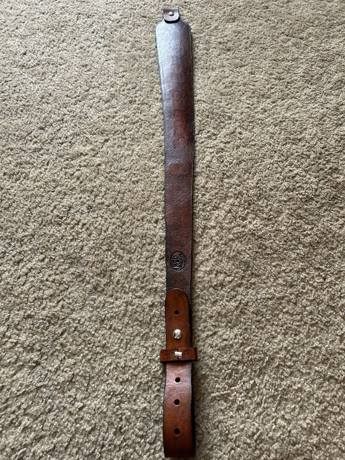 Brown leather leather rifle sling with smooth finish and brass screw holes, resting on a beige carpet.