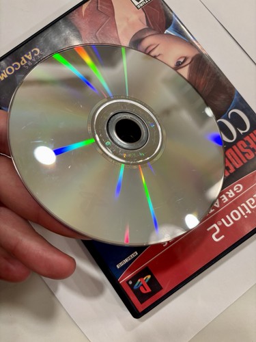 Hand holding a Resident Evil Code Veronica PS2 game disc with rainbow-colored dots and black label reading “Greatest Hits.”