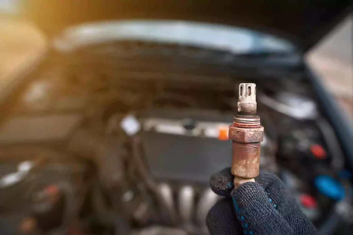 A mechanic wearing a black glove holding an old faulty oxygen sensor, with the vehicle’s engine in the background.