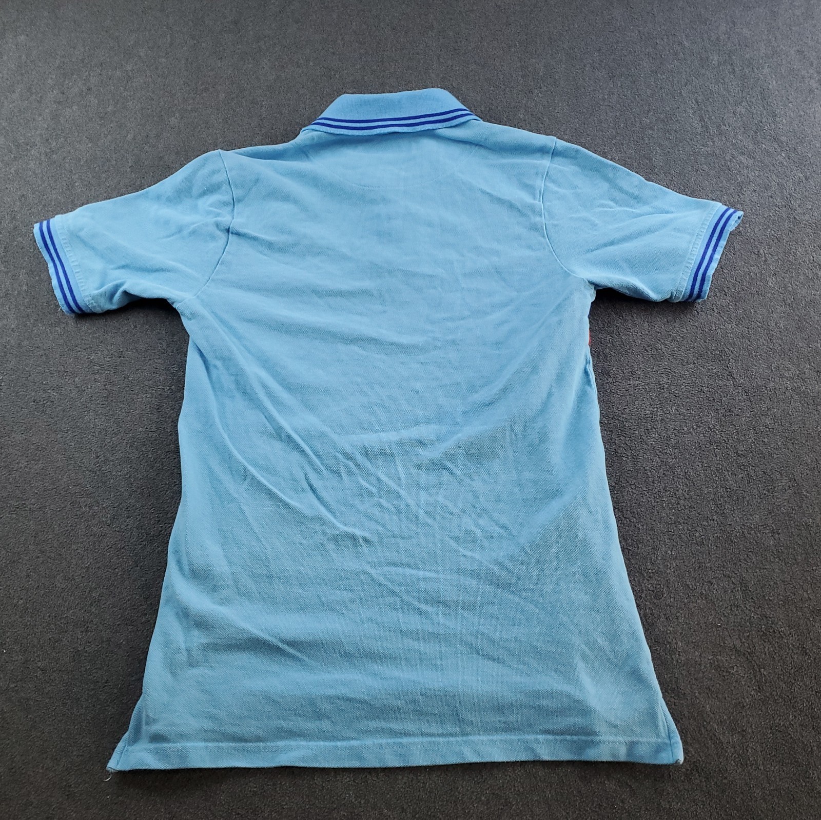 Light blue boys’ polo shirt with white sleeve trim, buttoned at the back, and blue Aston Martin Racing Team logo on chest.