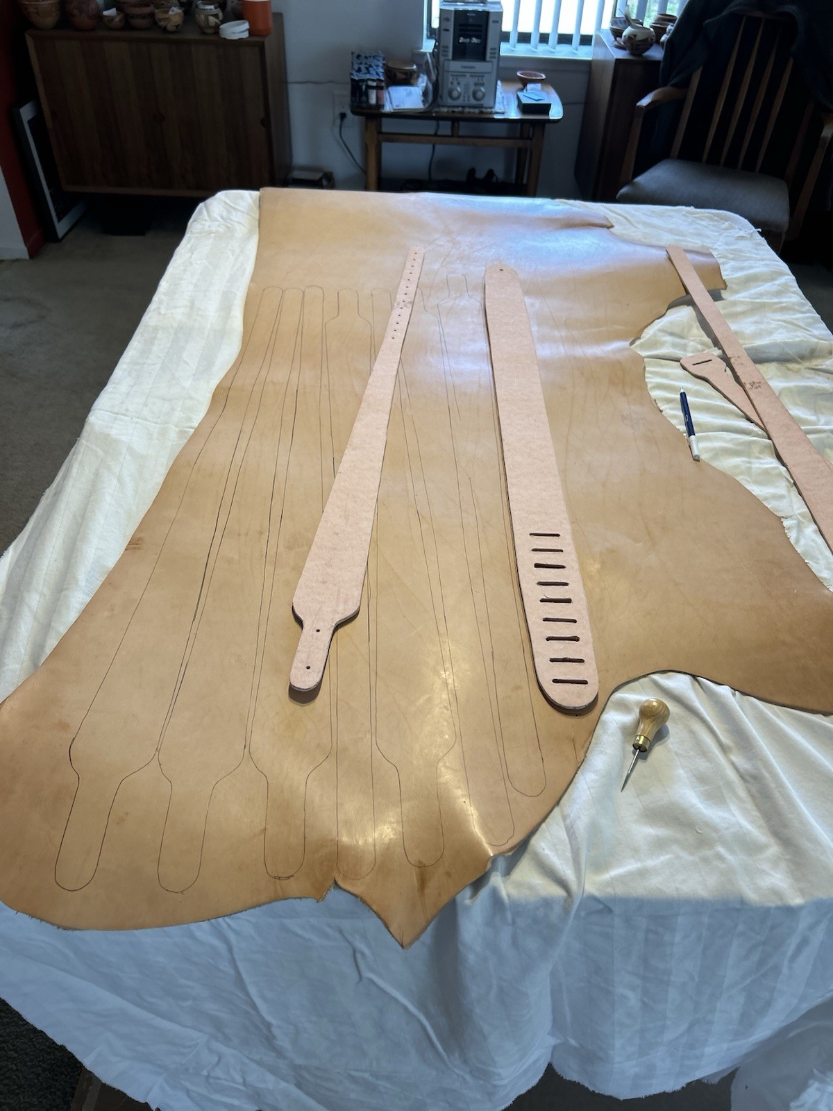 Brown leather rifle sling with two long handles and a wooden base on a white fabric table in a workshop setting.