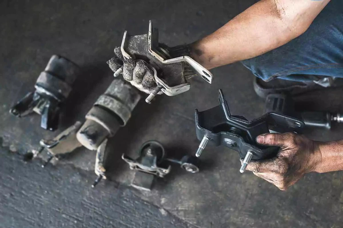A mechanic compares two engine mounts, with an old engine mount in his right hand and a new engine mount in his left hand. He is wearing a knit work glove on his right hand.