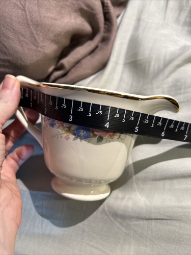 Cream ceramic mug with gold rim, floral print, and two measurement markings in centimeters on a light fabric surface.