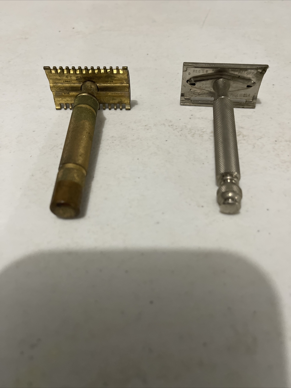Two vintage Gillette safety razors, one brass with flat rectangular head and one silver with flat rectangular head and black handle.